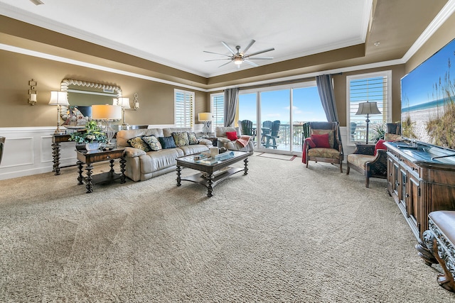 carpeted living room with a raised ceiling, wainscoting, ceiling fan, crown molding, and a decorative wall