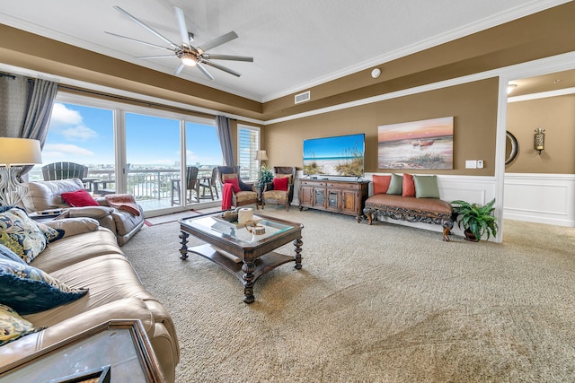 carpeted living area with visible vents, a ceiling fan, wainscoting, crown molding, and a decorative wall