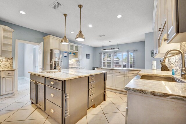 kitchen with hanging light fixtures, a center island with sink, tasteful backsplash, stainless steel fridge, and light tile floors