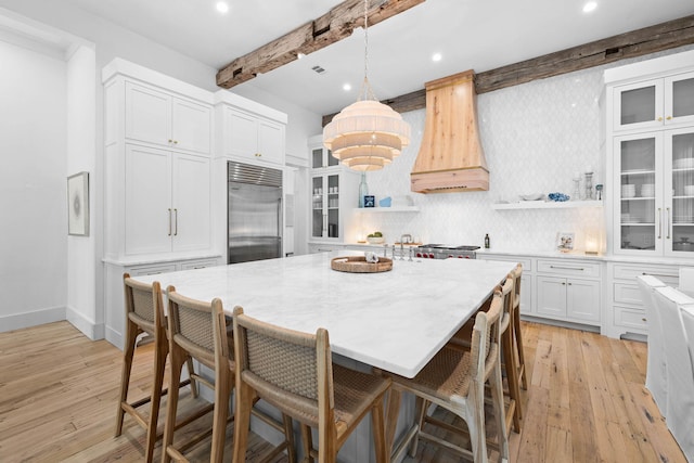 kitchen featuring premium range hood, light hardwood / wood-style flooring, a breakfast bar area, built in fridge, and beamed ceiling