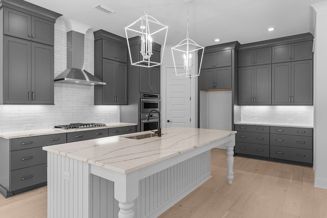 kitchen featuring tasteful backsplash, a kitchen island with sink, wall chimney range hood, light wood-type flooring, and appliances with stainless steel finishes