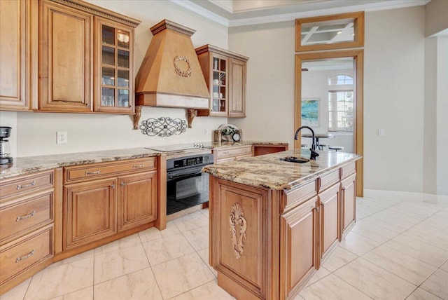 kitchen with premium range hood, black appliances, sink, a kitchen island with sink, and light stone countertops