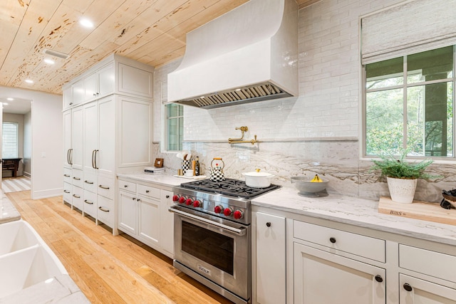 kitchen featuring a healthy amount of sunlight, designer stove, white cabinetry, and custom range hood