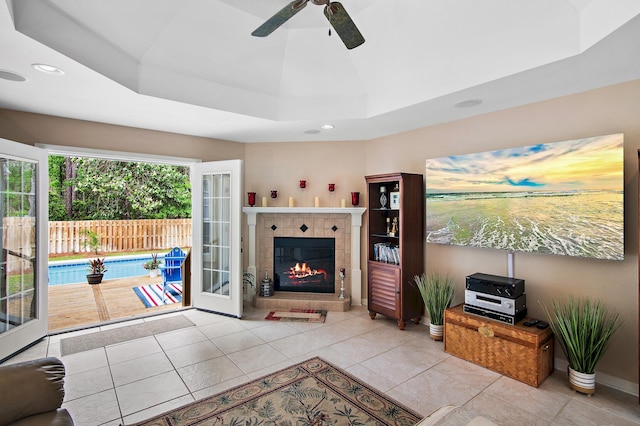 tiled living room with ceiling fan, a tray ceiling, and a fireplace