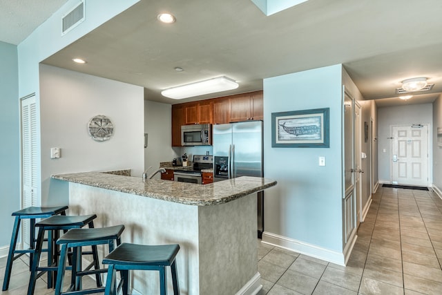 kitchen with light tile floors, a breakfast bar, stainless steel appliances, kitchen peninsula, and dark stone countertops