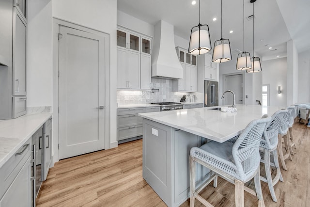 kitchen featuring premium range hood, light hardwood / wood-style flooring, hanging light fixtures, a center island with sink, and tasteful backsplash