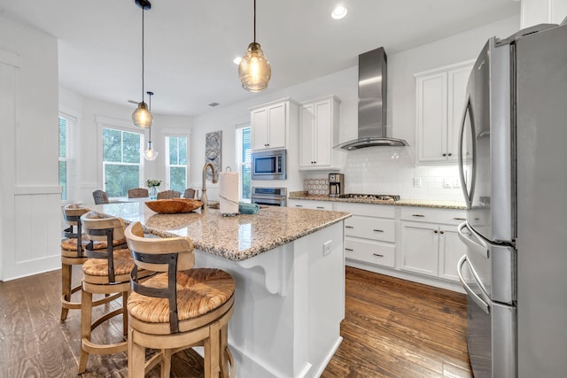 kitchen featuring hanging light fixtures, stainless steel appliances, an island with sink, wall chimney exhaust hood, and white cabinets