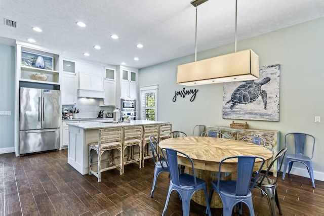 kitchen featuring dark wood-type flooring, decorative light fixtures, a center island with sink, stainless steel appliances, and white cabinets