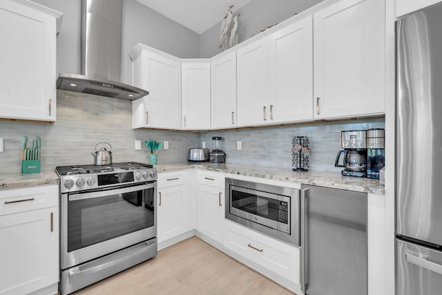 kitchen with appliances with stainless steel finishes, white cabinets, wall chimney range hood, backsplash, and light hardwood / wood-style flooring