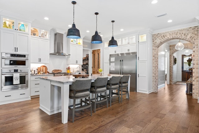 kitchen with wall chimney exhaust hood, dark wood-type flooring, appliances with stainless steel finishes, a kitchen bar, and a center island with sink