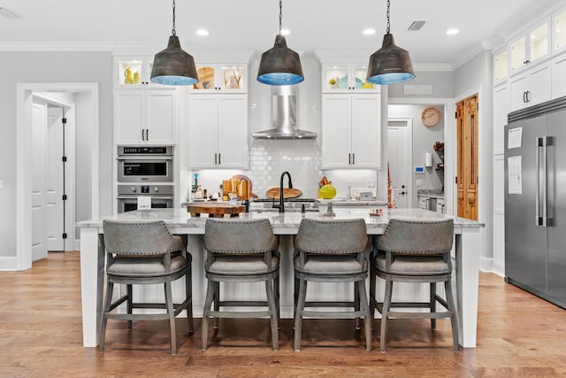 kitchen with wall chimney exhaust hood, appliances with stainless steel finishes, light hardwood / wood-style flooring, and a center island with sink