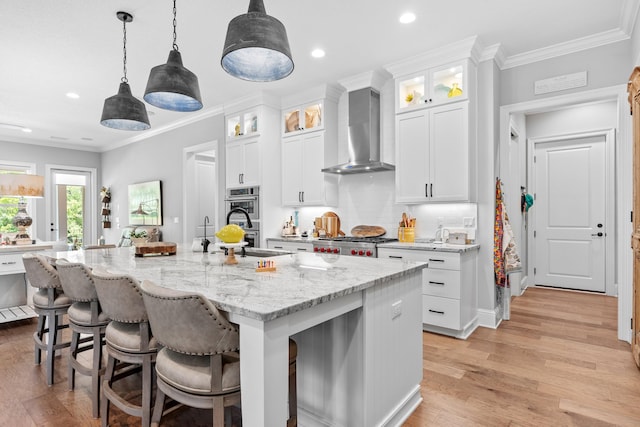 kitchen featuring wall chimney exhaust hood, light wood-type flooring, crown molding, a center island with sink, and pendant lighting