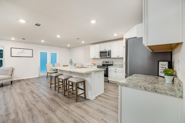 kitchen featuring appliances with stainless steel finishes, light stone counters, a center island with sink, light hardwood / wood-style floors, and white cabinetry