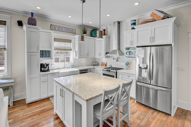 kitchen with wall chimney exhaust hood, hanging light fixtures, stainless steel appliances, a kitchen island, and light hardwood / wood-style floors