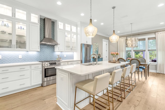 kitchen with wall chimney range hood, a center island with sink, light hardwood / wood-style flooring, sink, and stainless steel appliances