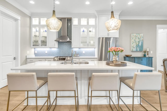 kitchen with wall chimney range hood, stainless steel fridge, a center island with sink, white cabinetry, and light wood-type flooring