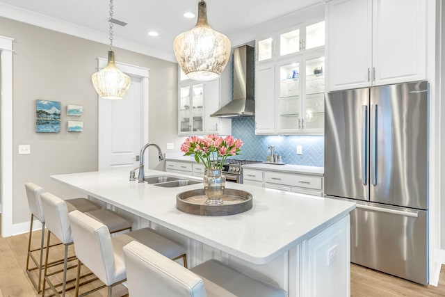 kitchen with wall chimney range hood, hanging light fixtures, an island with sink, appliances with stainless steel finishes, and light hardwood / wood-style flooring