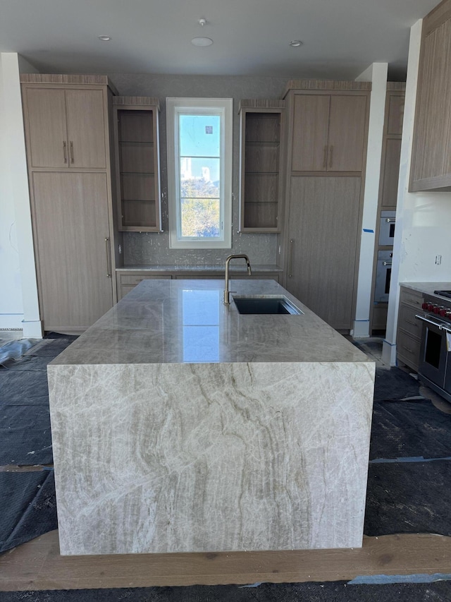 kitchen with light stone counters, stainless steel appliances, light brown cabinets, a sink, and an island with sink