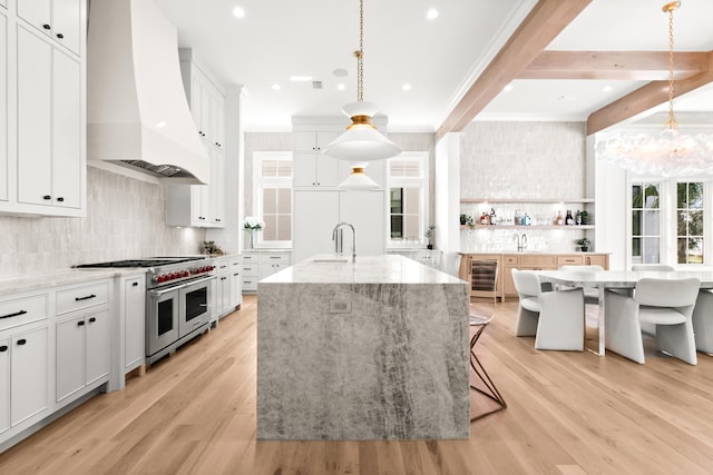 kitchen featuring white cabinetry, range with two ovens, custom exhaust hood, a kitchen island with sink, and light wood-type flooring