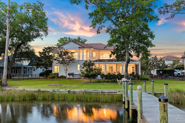 back house at dusk featuring a water view, a balcony, and a lawn