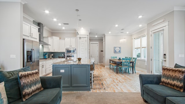 kitchen with wall chimney exhaust hood, white cabinets, light stone countertops, stainless steel appliances, and an island with sink