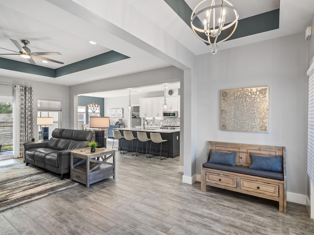 living room with sink, ceiling fan with notable chandelier, light wood-type flooring, and a tray ceiling