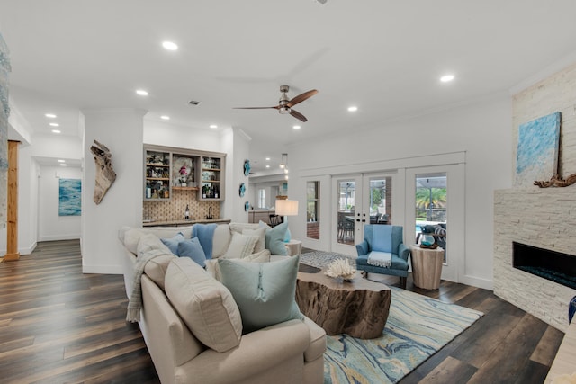 living room with ceiling fan, a stone fireplace, dark wood-type flooring, crown molding, and french doors