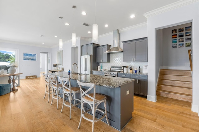 kitchen with wall chimney exhaust hood, premium appliances, light hardwood / wood-style floors, ornamental molding, and a center island with sink