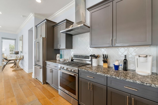 kitchen with wall chimney range hood, stainless steel appliances, tasteful backsplash, dark stone countertops, and light hardwood / wood-style floors