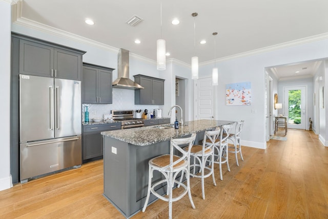 kitchen featuring wall chimney exhaust hood, a kitchen island with sink, decorative light fixtures, premium appliances, and light hardwood / wood-style floors