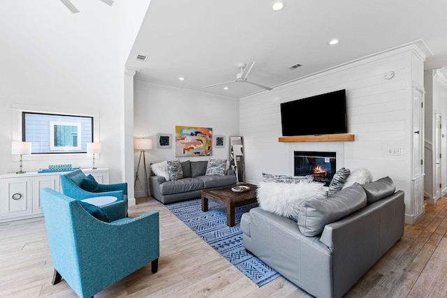 living area with light wood finished floors, visible vents, ornamental molding, a glass covered fireplace, and a ceiling fan