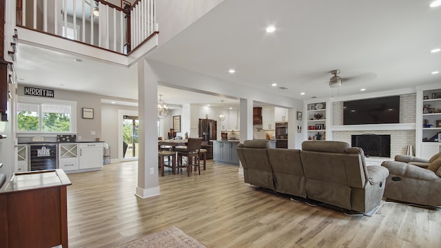 living room with ceiling fan with notable chandelier, wine cooler, a brick fireplace, built in shelves, and light hardwood / wood-style floors