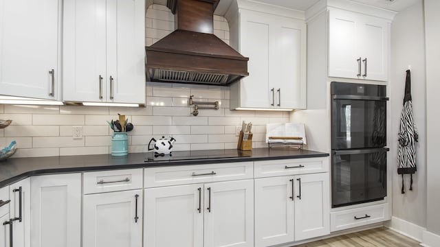 kitchen with backsplash, white cabinetry, black double oven, and custom exhaust hood