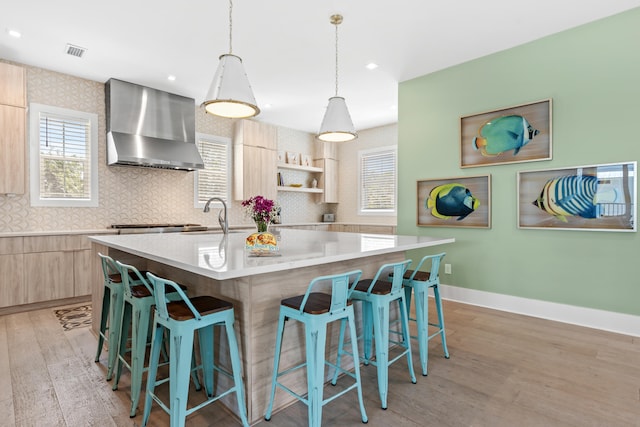 kitchen with an island with sink, wall chimney exhaust hood, light hardwood / wood-style floors, and light brown cabinetry