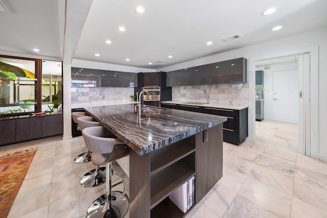 kitchen with sink, tasteful backsplash, dark stone countertops, a kitchen bar, and a center island with sink