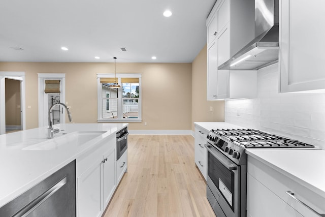 kitchen featuring light wood-type flooring, stainless steel appliances, wall chimney range hood, white cabinets, and hanging light fixtures