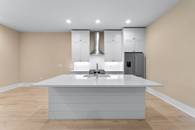 kitchen featuring wall chimney exhaust hood, sink, white cabinets, stainless steel fridge with ice dispenser, and an island with sink