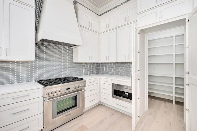 kitchen featuring white cabinetry, premium range hood, light hardwood / wood-style floors, decorative backsplash, and appliances with stainless steel finishes