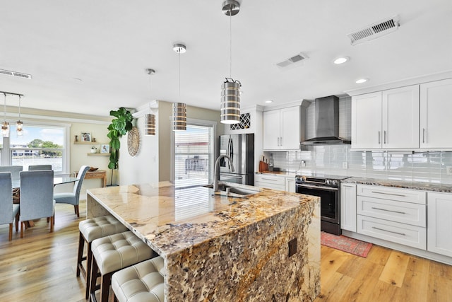 kitchen featuring white cabinetry, wall chimney range hood, an island with sink, pendant lighting, and appliances with stainless steel finishes