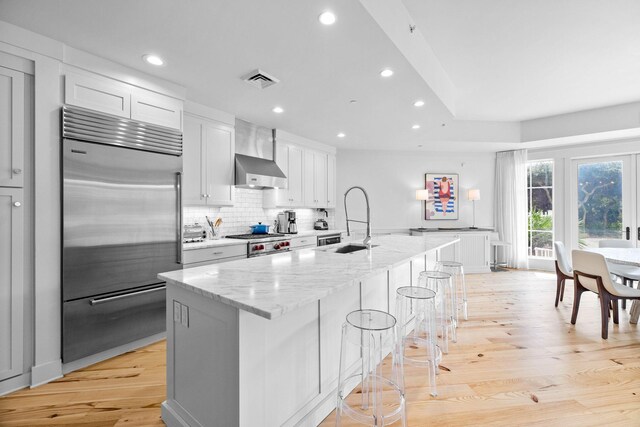 kitchen with a sink, stainless steel built in fridge, wall chimney exhaust hood, light wood-type flooring, and backsplash