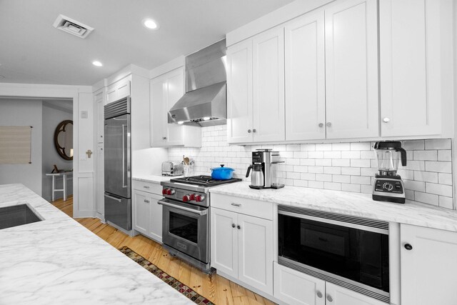 kitchen featuring light wood finished floors, visible vents, wall chimney range hood, light stone countertops, and built in appliances