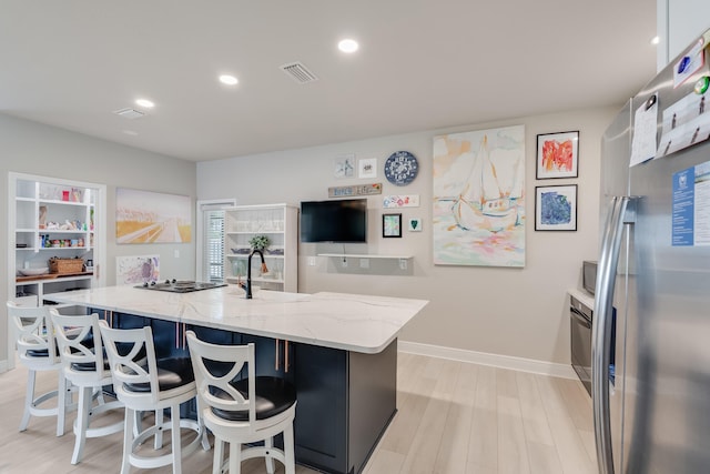 kitchen with sink, an island with sink, light hardwood / wood-style floors, light stone counters, and stainless steel refrigerator