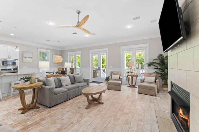 living room featuring crown molding, light hardwood / wood-style flooring, a tile fireplace, ceiling fan, and french doors