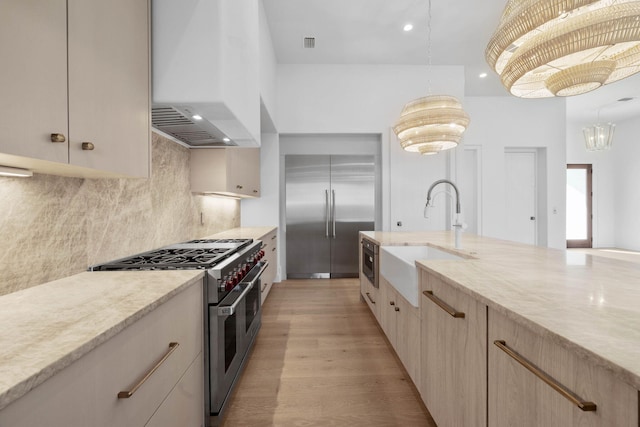 kitchen featuring high quality appliances, wall chimney range hood, sink, hanging light fixtures, and light hardwood / wood-style flooring