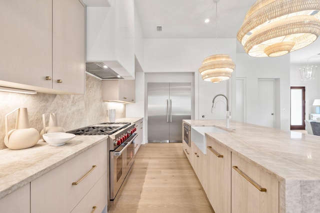 kitchen with a sink, high quality appliances, wall chimney range hood, light wood-type flooring, and decorative backsplash