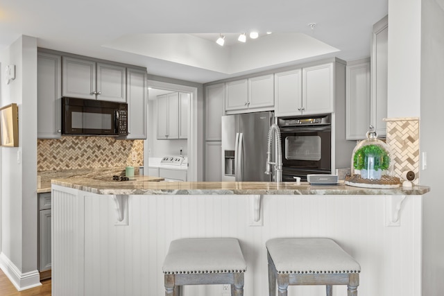 kitchen featuring kitchen peninsula, a kitchen bar, gray cabinetry, washer and clothes dryer, and black appliances