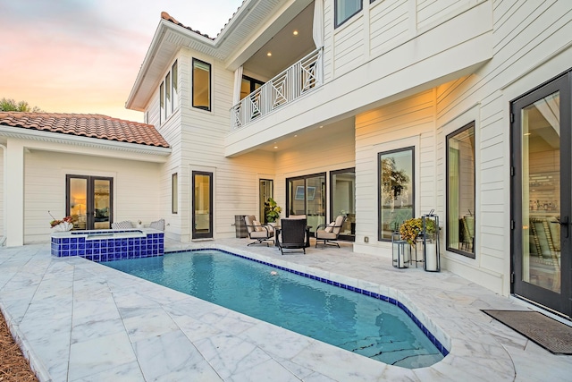 back house at dusk featuring a swimming pool with hot tub, a balcony, an outdoor hangout area, and a patio