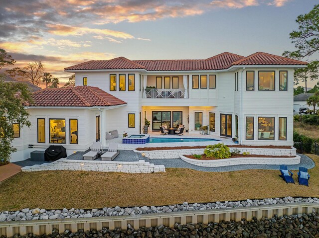 back house at dusk featuring an outdoor living space, a swimming pool with hot tub, a balcony, a yard, and a patio area