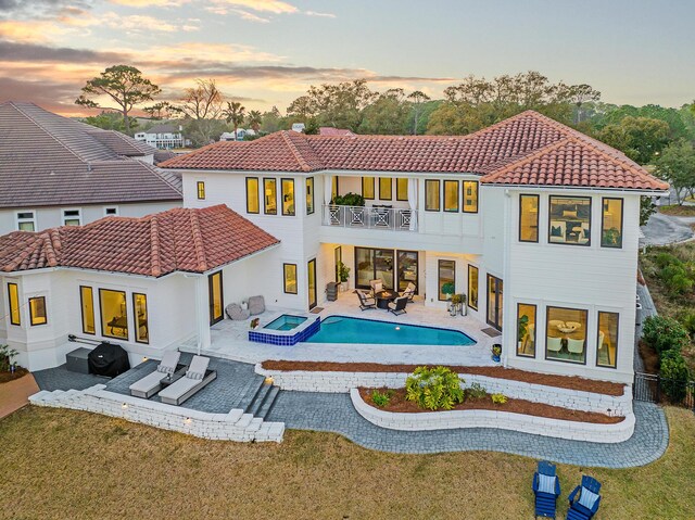 back house at dusk with a yard, a balcony, a patio area, and a pool with hot tub