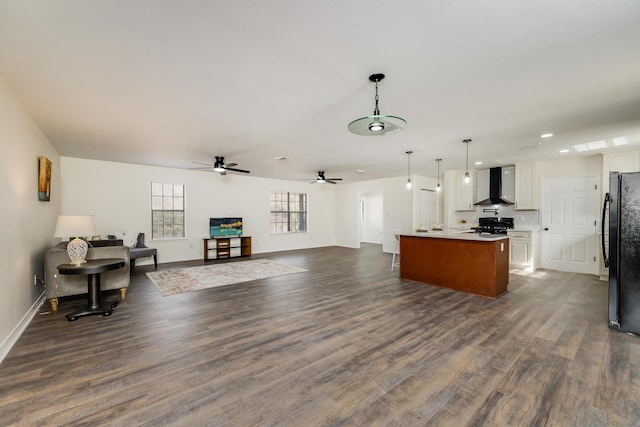 kitchen with dark wood-type flooring, black refrigerator, an island with sink, white cabinets, and wall chimney exhaust hood